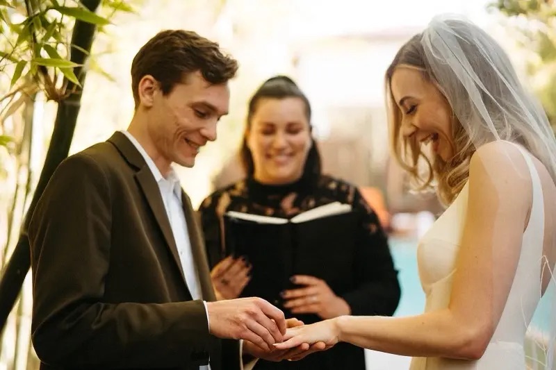 Groom sliding a ring onto his bride’s finger while the officiant smiles beside them