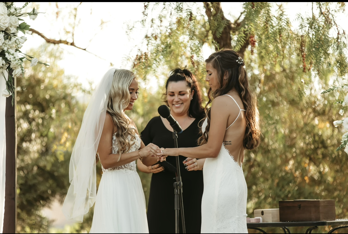 Two brides smiling and holding hands as they exchange vows with officiant nearby